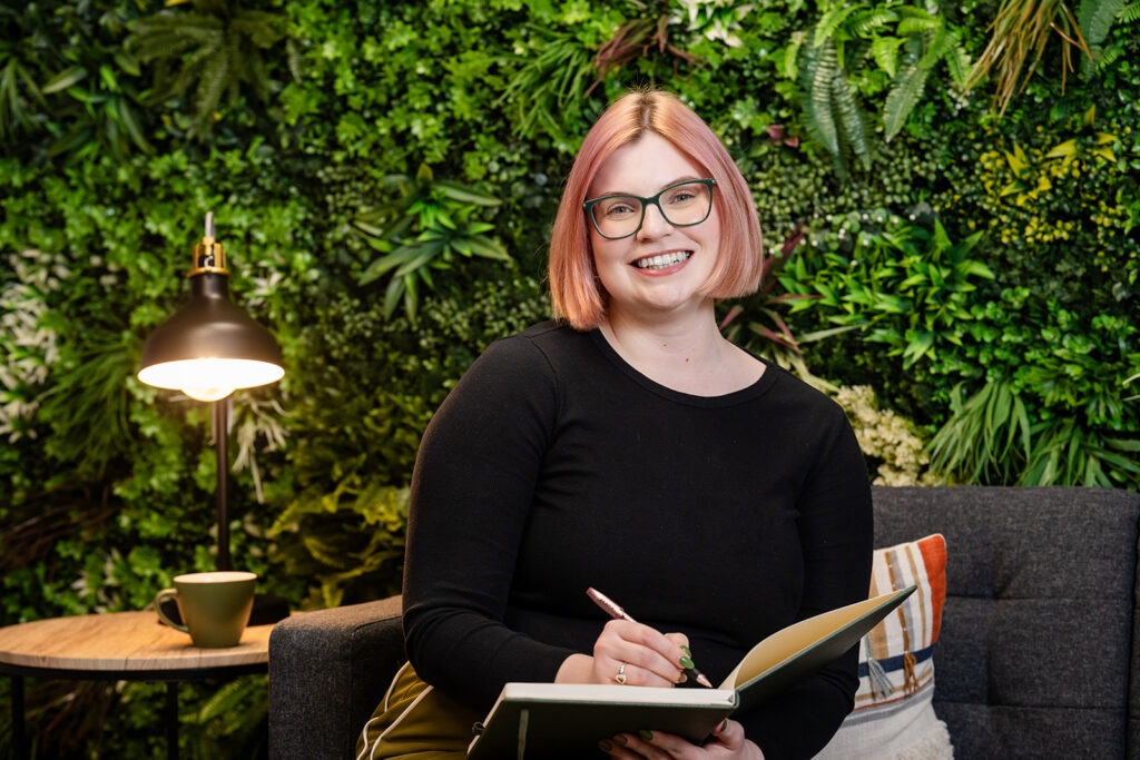 Dr Charlotte Davies sitting with a book beside a table with a lamp on it.
